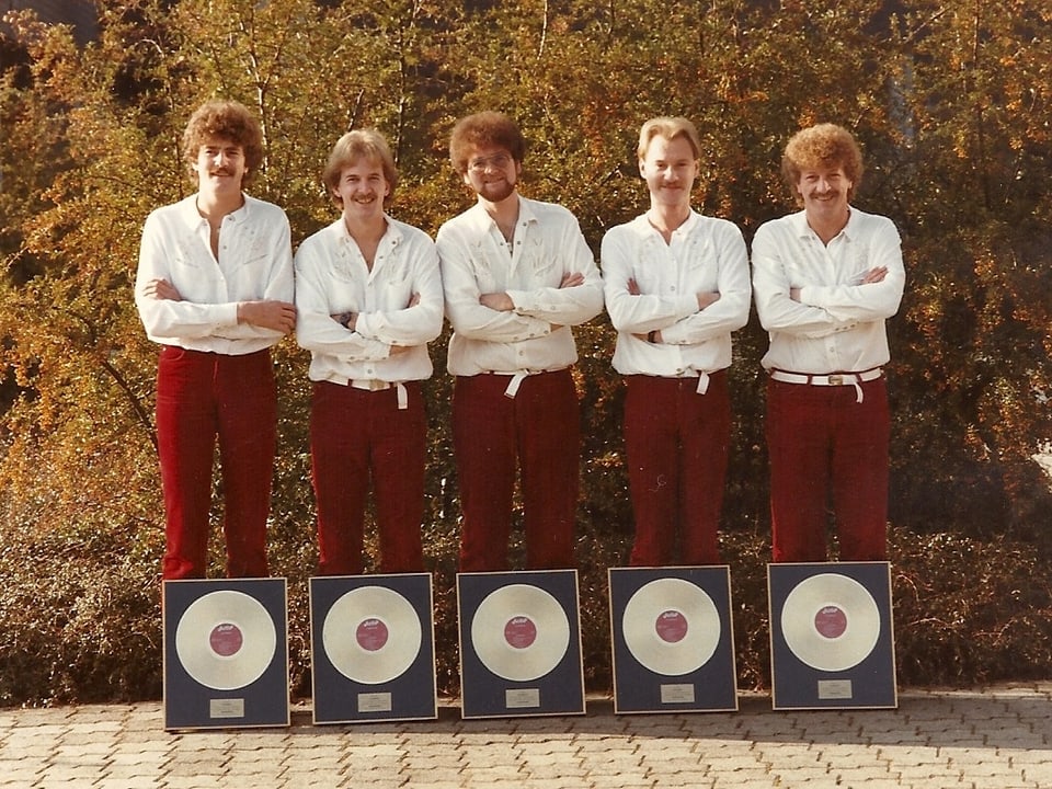 Fünf Männer mit weissen Hemden und roten Hosen stehen vor fünf goldenen Schallplatten.