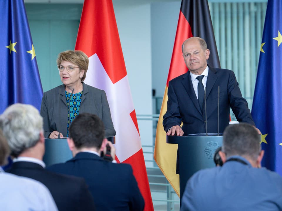 Zwei Politiker bei einer Pressekonferenz, mit den Flaggen der EU, Schweiz und Deutschland im Hintergrund.