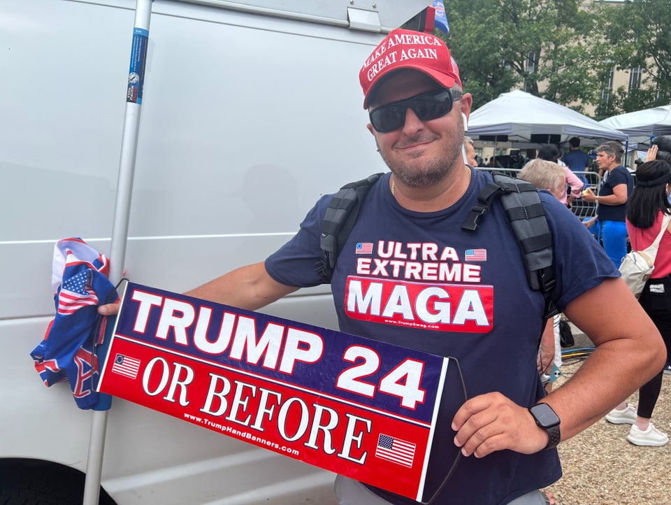 Ein Mann trägt ein T-Shirt und ein Schild, die beide Werbung für Donald Trump machen.