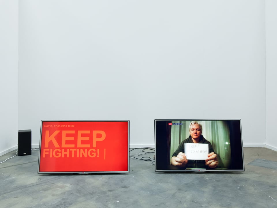 Zwei Bildschirme stehen nebeneinander am Boden. Auf dem linken steht "Keep Fighting", auf dem rechten ist Julian Assange zu sehen.