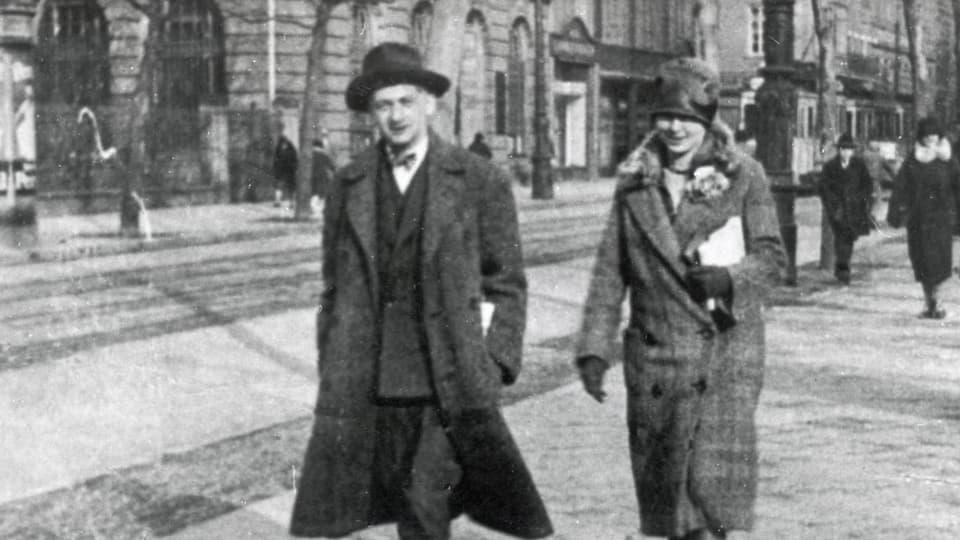 schwarzweiss Foto Mann in langem Mantel und Hut, daneben Frau mit Hut, auf den Strassen, dahinter ein altes Tram