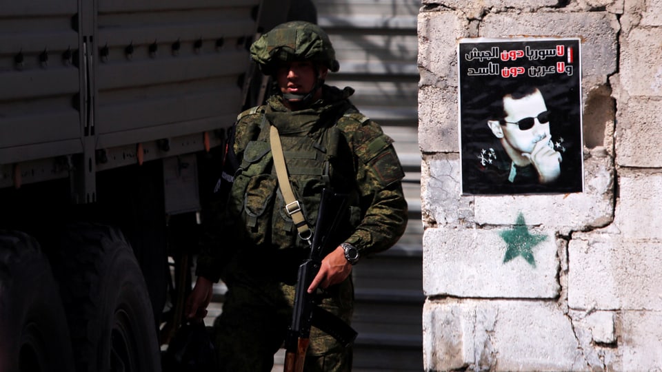 Bewaffneter Soldat steht neben einer Mauer, auf der ein Plakat mit dem Konterfei von Baschar al-Asssad hängt.