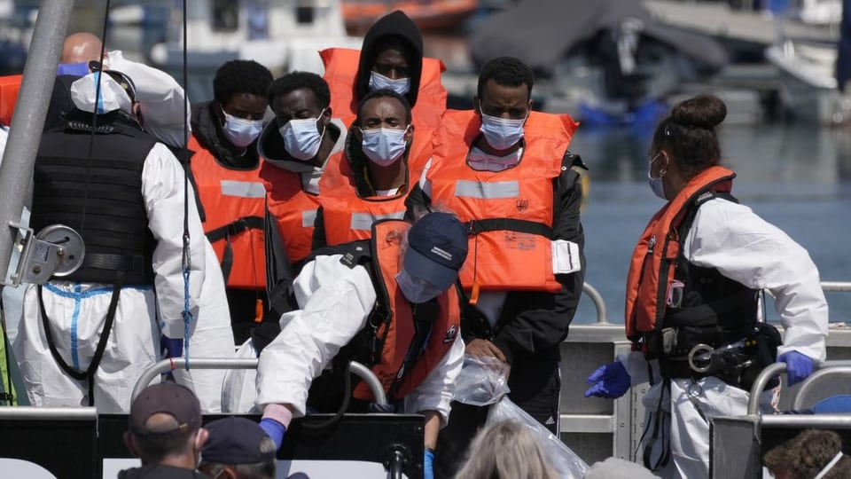 Mehrere Migranten stehen orangen Schwimmwesten auf einem Boot