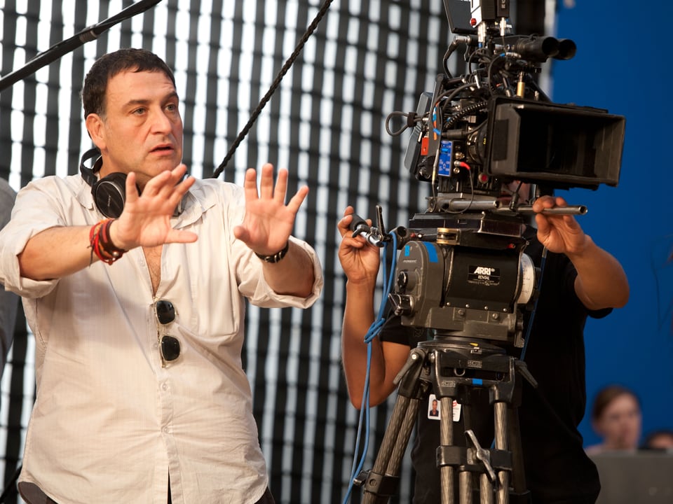 Regisseur in weissem Hemd steht neben der Kamera, Hände in der Luft.