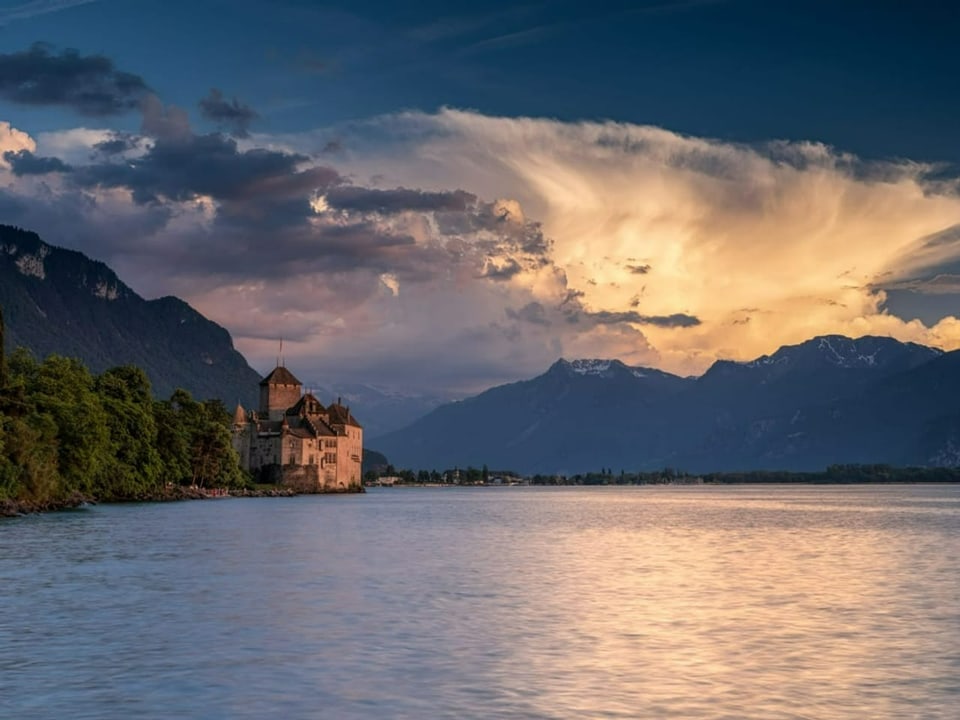 Blick vom See auf ein Schloss am linken Ufer. Rechts hinter Bergen imposante Gewitterwolke, die im Abendlicht leichtet.