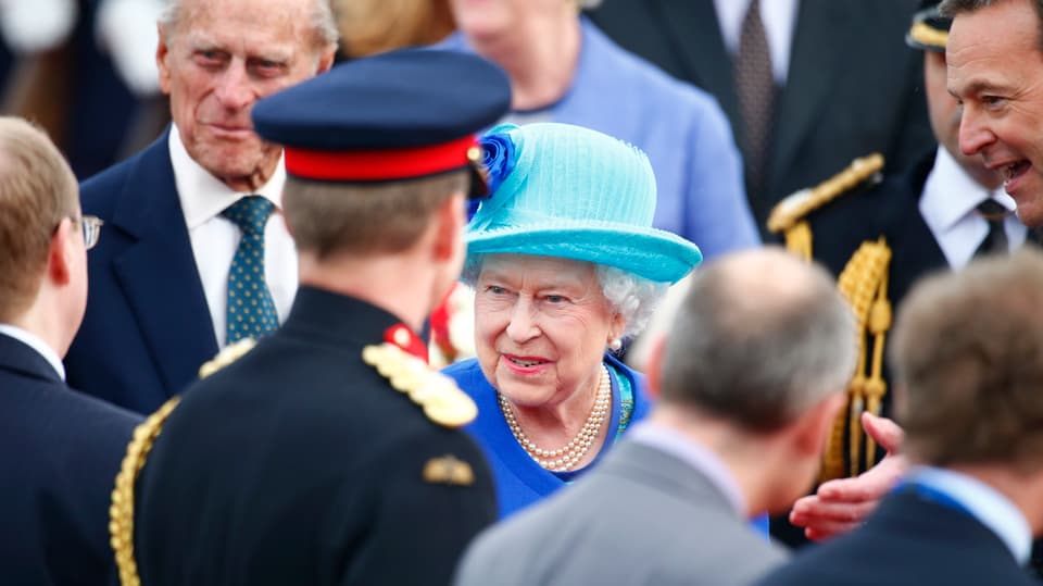 Queen Elizabeth II. in der Mitte des Bildes mit blauem Hut in einer Menschenmenge stehend
