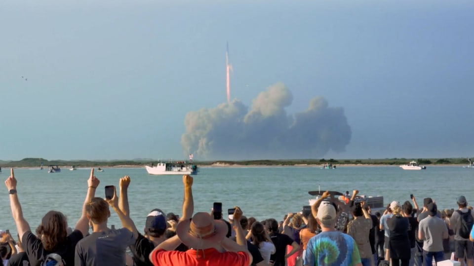 Menschenmenge mit Rücken zur Kamera schaut aufs Meer, eine Rakete steigt in Rauchwolke auf.