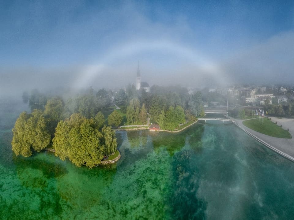 Luftfoto vom Ufer an einem See mit Nebelschwaden. In der Mitte ein kreisförmiger weisser Bogen. 