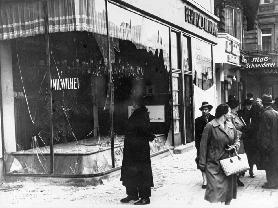 Passanten gehen an einer zerstoerten Fensterfront eines juedischen Geschaefts vorbei.