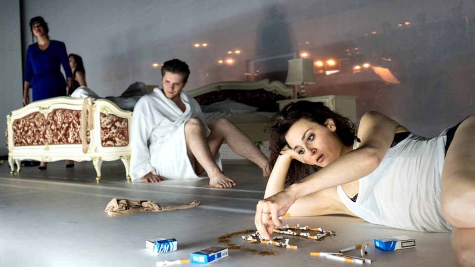 Frau liegt auf Boden und spielt mit Zigaretten, Mann lehnt an Bett.