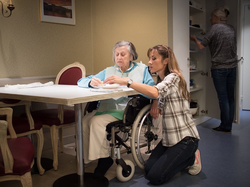 Corinne Waldmeier kniet neben eine im Rollstuhl sitzenden Bewohnerin an einem Tisch.
