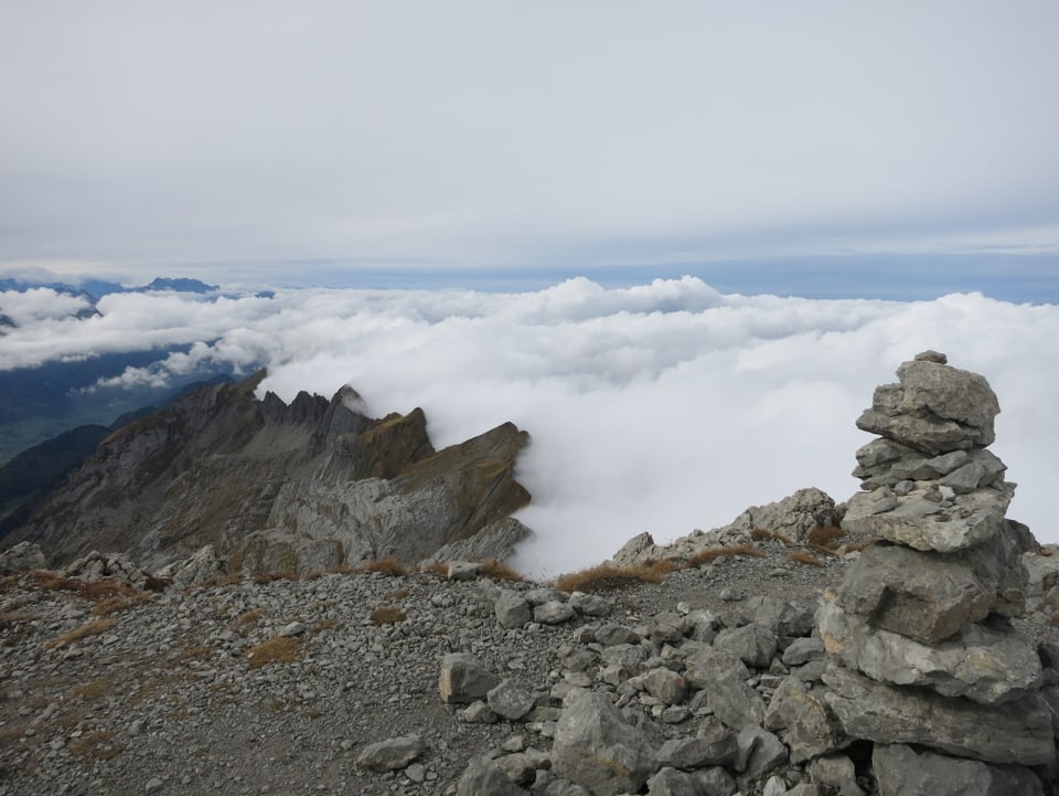 Ein Steinmännchen steht im Vordergrund des Bildes, der Betrachter befindet auf dem Gipfel des Girenspitz in der Alpsteinregion. Ein Gebirgskamm zieht quer durch das Bild. Links des Kammes ist die Luft klar, rechts des Kammes liegen viele Wolken.