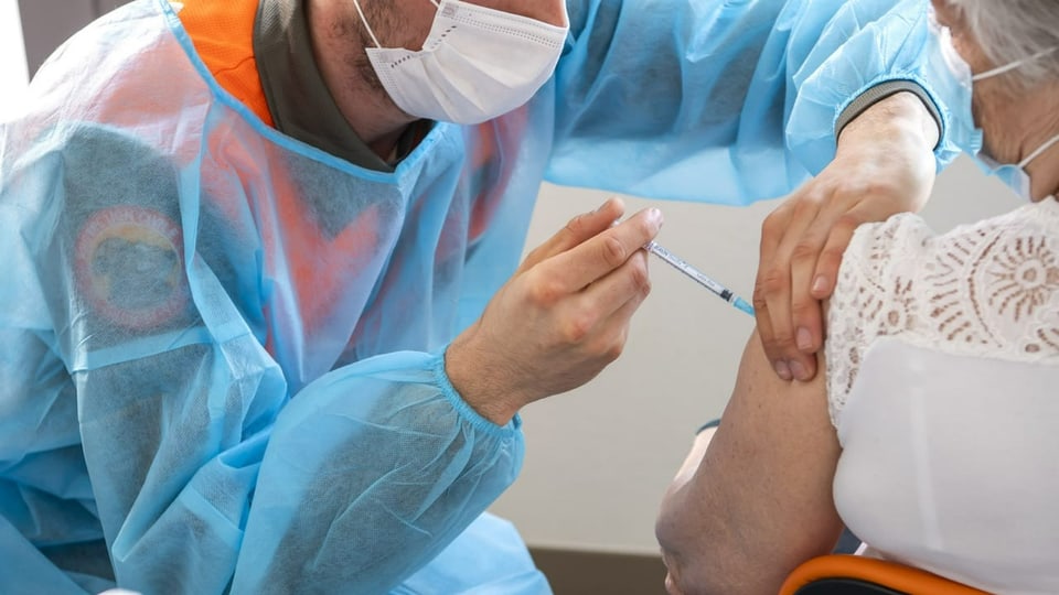Ein Mann in blauem Schutzanzug setzt eine Impfung bei einer Frau.
