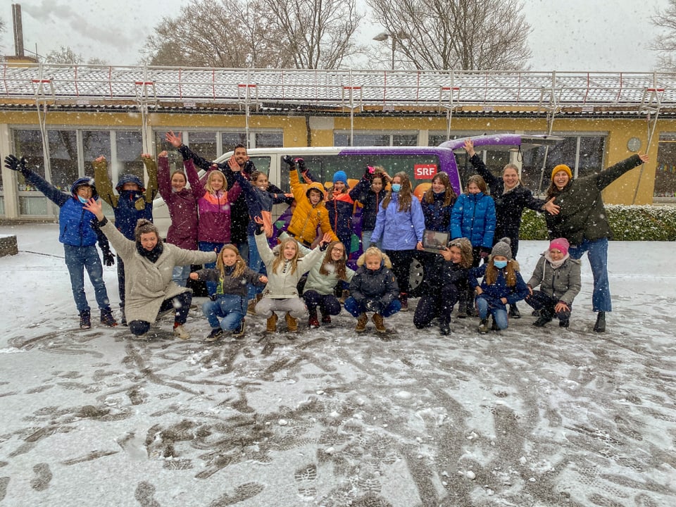 Die Klasse steht vor dem Zambobus und alle Personen strecken die Arme in die Luft. Es liegt Schnee und schneit.