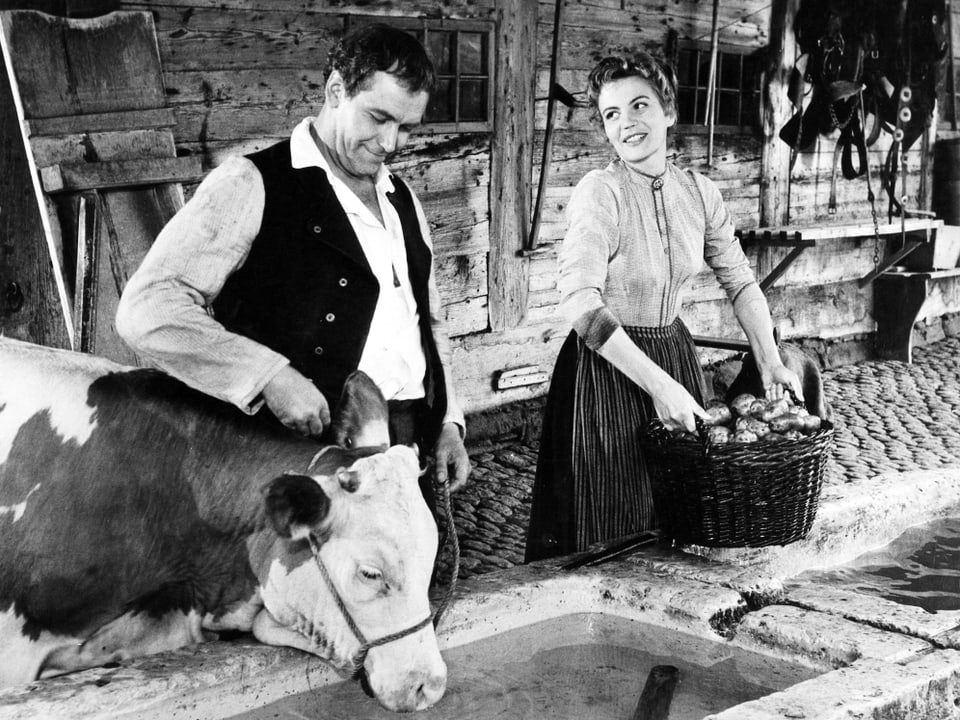 Ein Mann hält eine Kuh beim Halsband, die Kuh trinkt aus einem Trog. Neben dem Mann steht eine junge Frau mit einem Korb voll Kartoffeln.