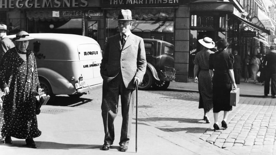 historische Schwarzweiss-Aufnahme eines alten, schlanken Mannes, der mit Anzug und Hut und Stock spazieren geht.