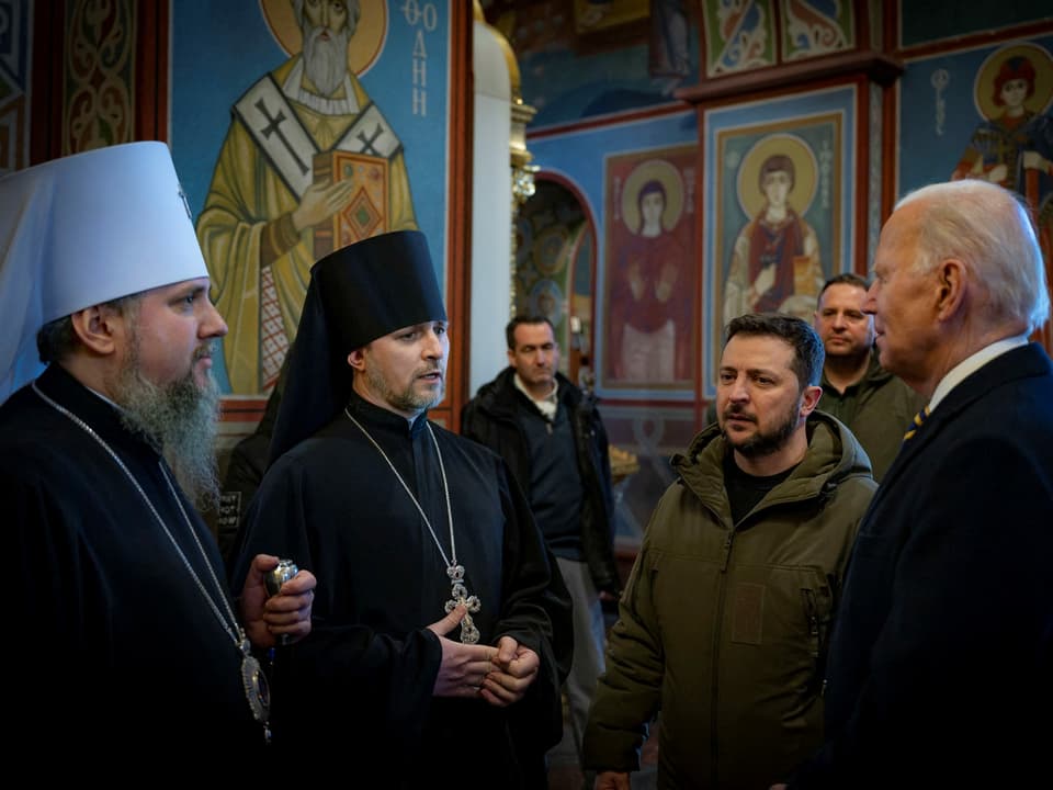 Vier Männer stehen zusammen, die zwei linken in traditioneller Kleidung der orthodoxen Kirche - jeweils einer mit weissem Hut, der andere mit schwarzem Hut.