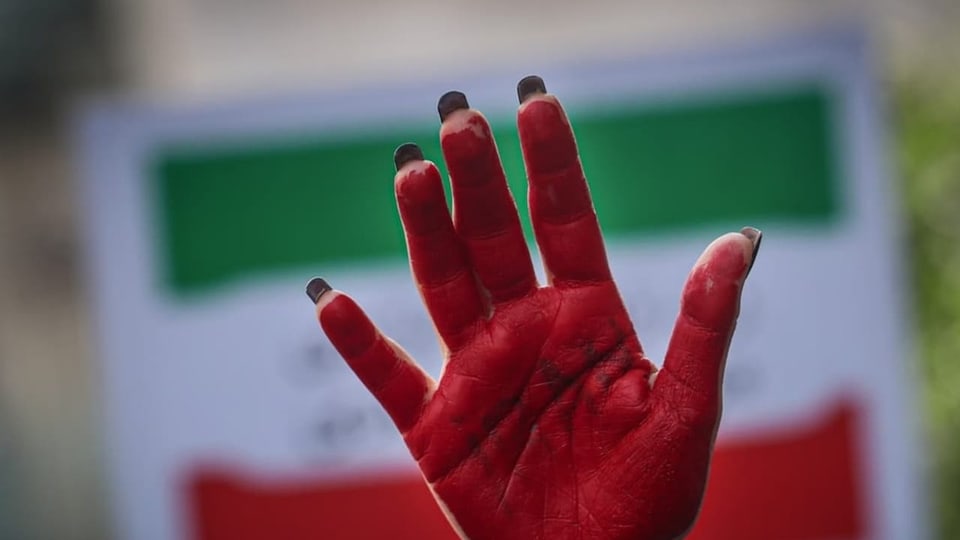 Eine Person hält eine rot bemalte Handfläche in die Luft. Im Hintergrund ist unscharf eine Iran-Flagge.