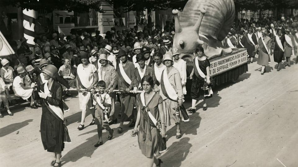 eine schwarz-weiss Fotografie von demonstrierenden Frauen, die eine grosse Schnecke ziehen