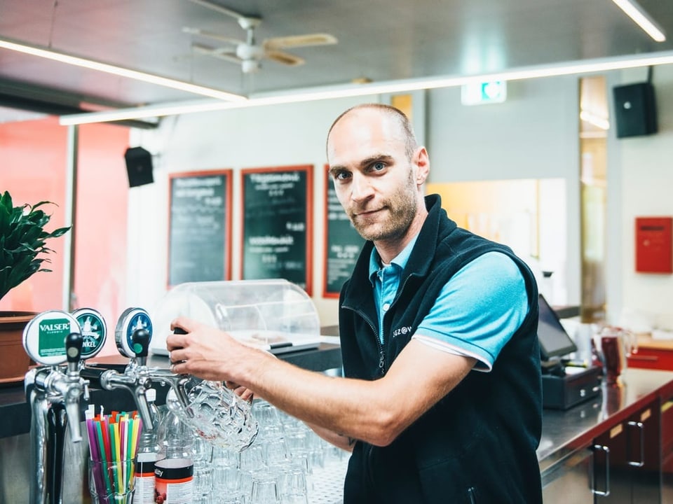 Ein Mann steht in einem Fleece-Gilet an einer Bar und zapft ein Glas Bier.