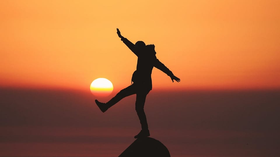 A person balances on a stone on their right leg. The sunset can be seen in the background.