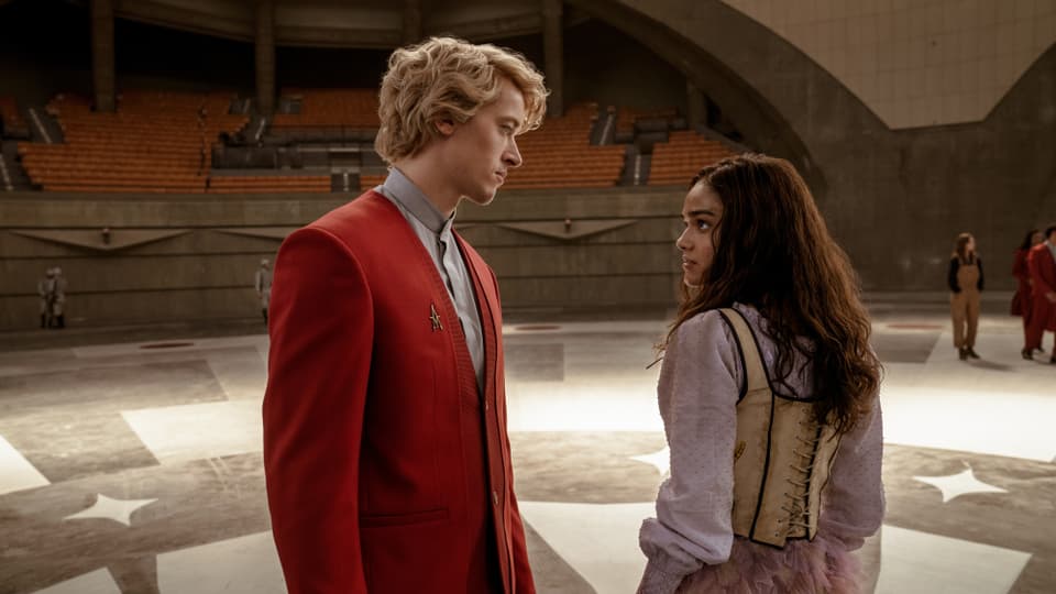A man in a red suit and blonde hair stands opposite a woman with black hair in a vest and dress.