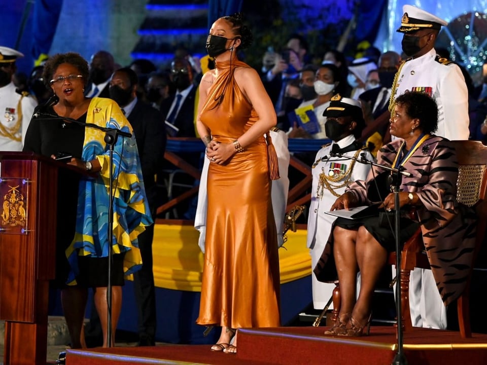 Rihanna steht mit geschlossenen Augen und Maske auf einem Podest, flankiert von Präsidentin und Premierministerin.