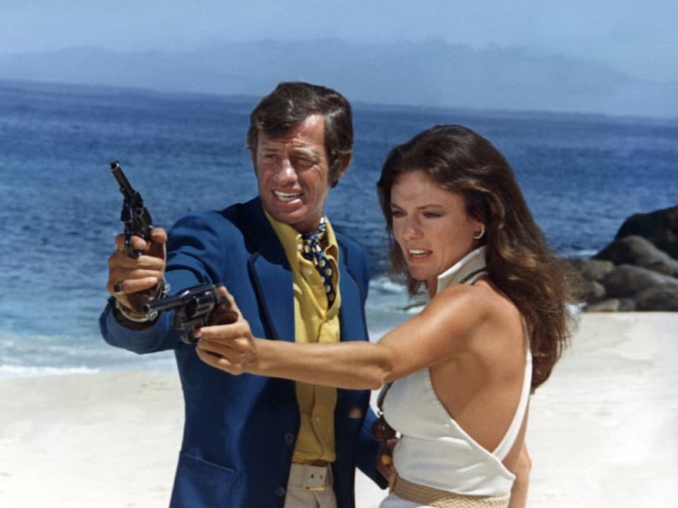 Eine Mann und eine Frau stehen am Strand, beide zücken eine Pistole und zielen.
