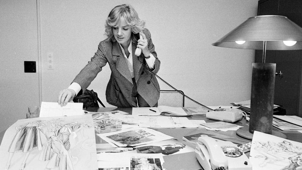 Eine jüngere blonde Frau steht mit Telefonhörer in der Hand hinter einem Schreibtisch, auf dem Zeichnungen liegen.