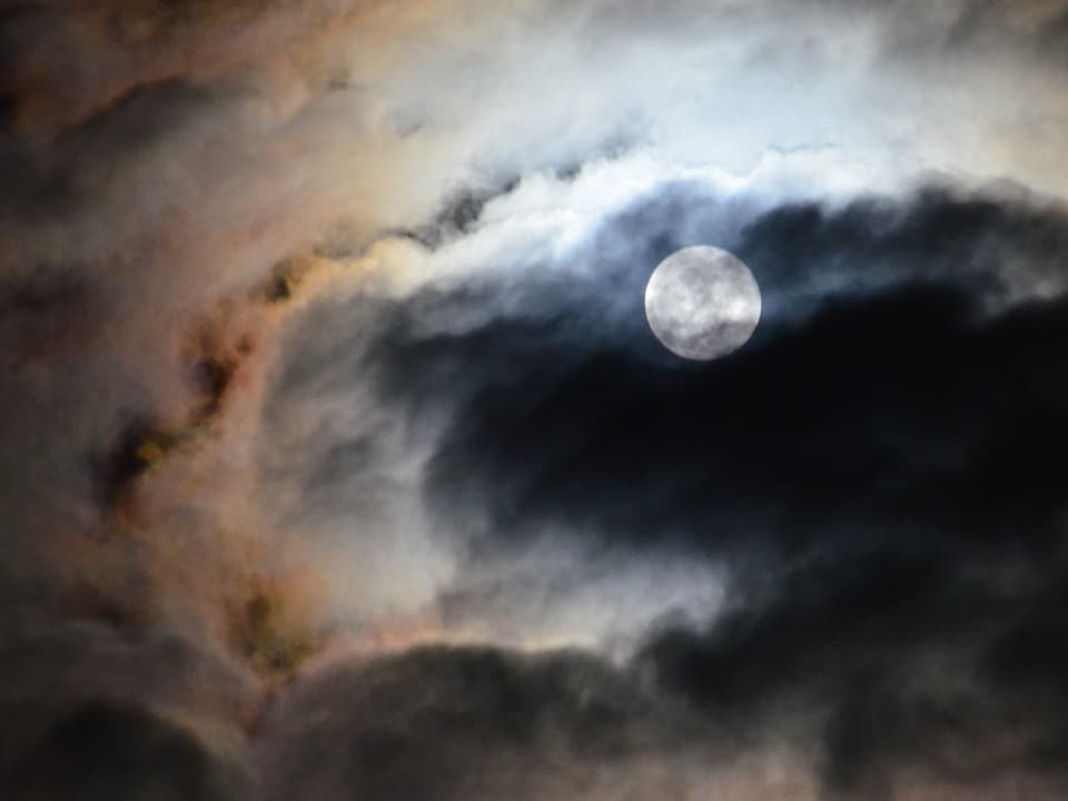 Wie durchgewirbelter Rauch zeigen sich die Wolken, dabei sind diese weiss, grau und gelb-rötlich gefärbt. Am Rand der Wolken ist der Mond sehr gut zu erkennen.
