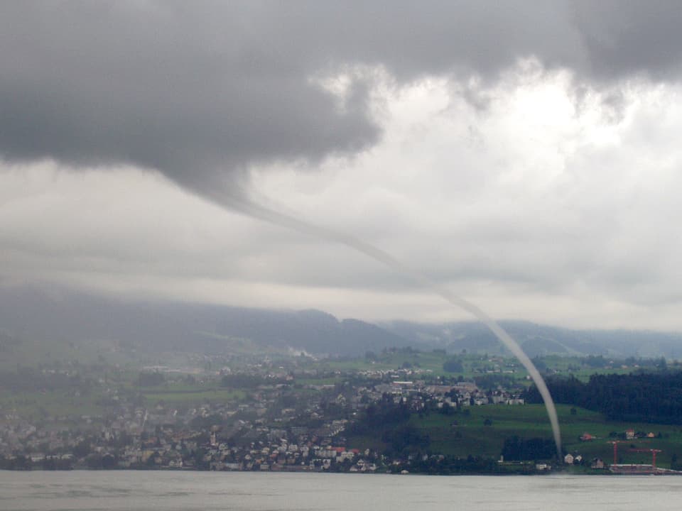 Ein grauer Wolkenrüssel senkt sich schräg von der grauen Wolke bis zum Zürichsee hinunter. Dahinter ist ein Dorf am Hang zu sehen.