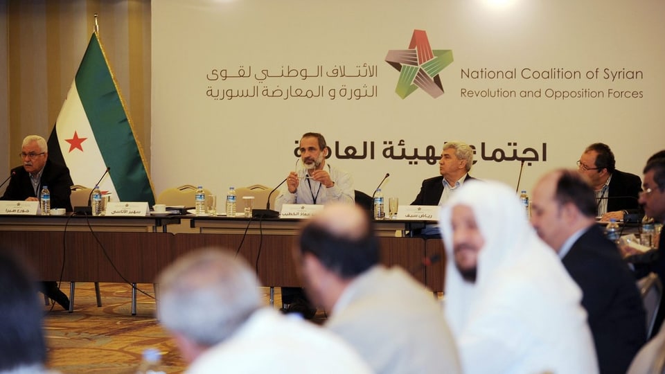 Vertreter der syrischen Opposition sitzen an einem Tisch, in der Mitte des Bildes ihr früherer Vorsitzender, Moaz Al-Kahtib.