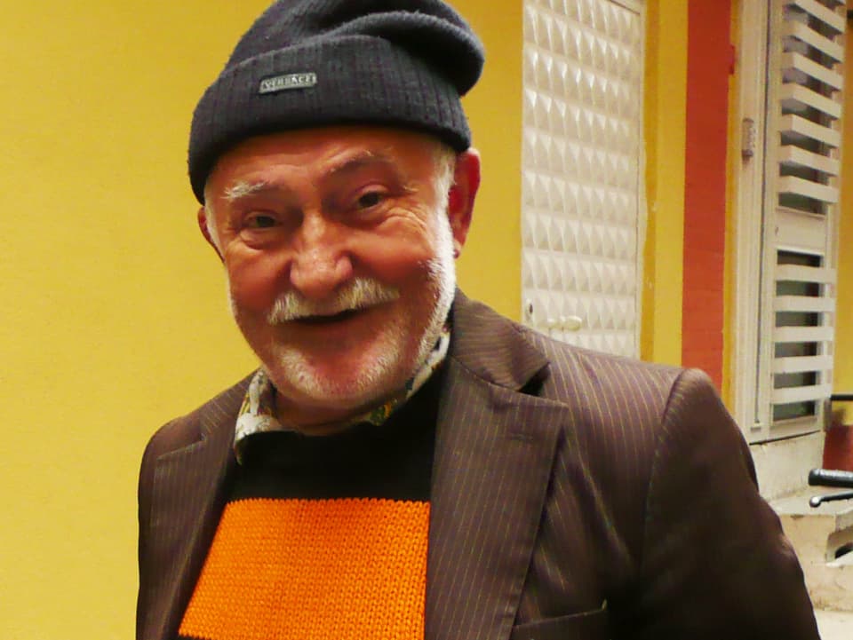 Ein älterer Mann mit Mütze und orange-schwarzem Pullover.