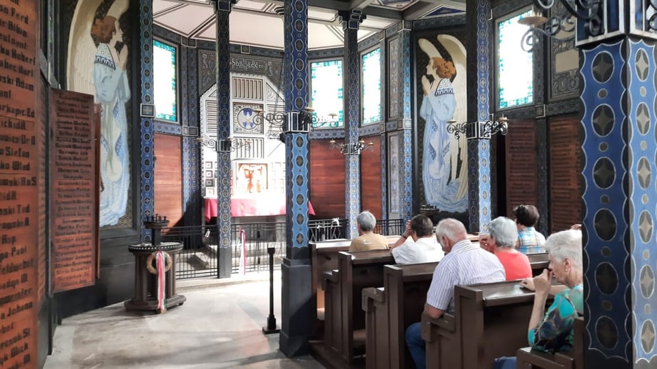 Innenraum einer Kapelle, rechts und links beschriebene Holzwände, in der Mitte Menschen auf Holzbänken, blaue Säulen.