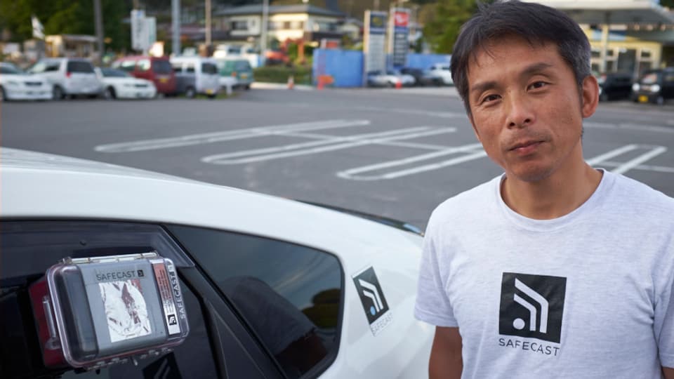 Auto mit Geigerzähler an der Scheibe, daneben ein Japaner um die vierzig mit SAFECAST T-Shirt-