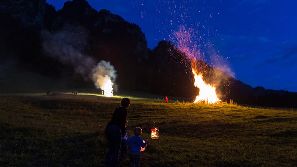 Ein Höhenfeuer auf einer Anhöhe im Churer Rheintal. Daneben brennt ein Vulkan. Im Vordergrund sitzen ein Erwachsener und ein Kind im Dunkeln.