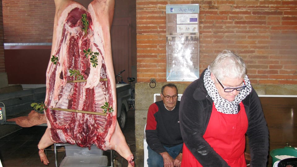 Ein Marktstand: Ein halbes Schwein hängt von der Decke, daneben steht eine Frau mit dickem Schal und roter Schürze.