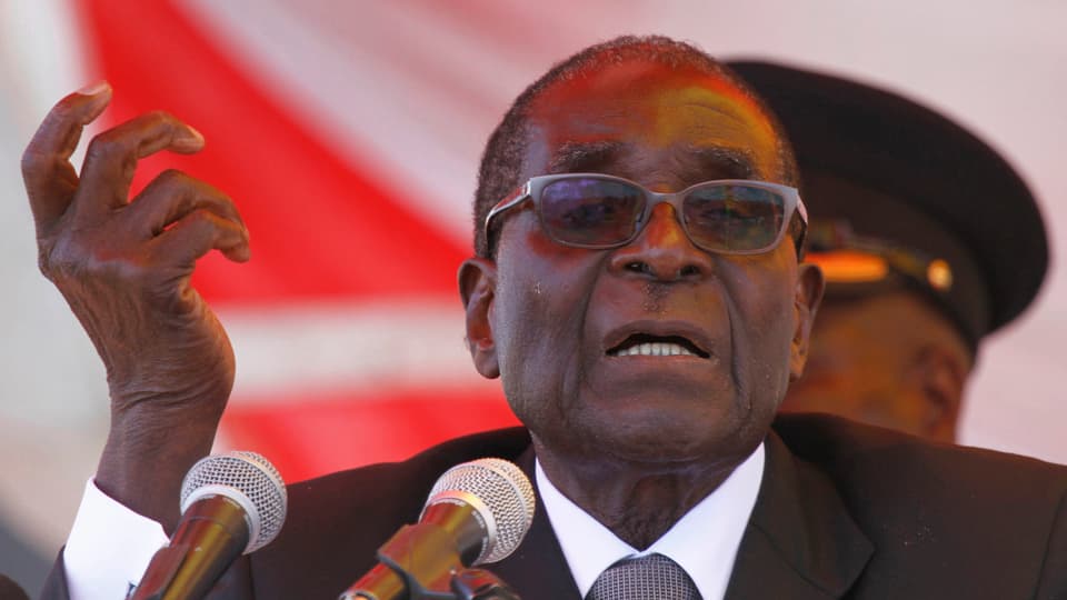 Mugabe bei einer Ansprache mit erhobener Hand, im Hintergrund ein Uniformträger und eine rote Fläche.