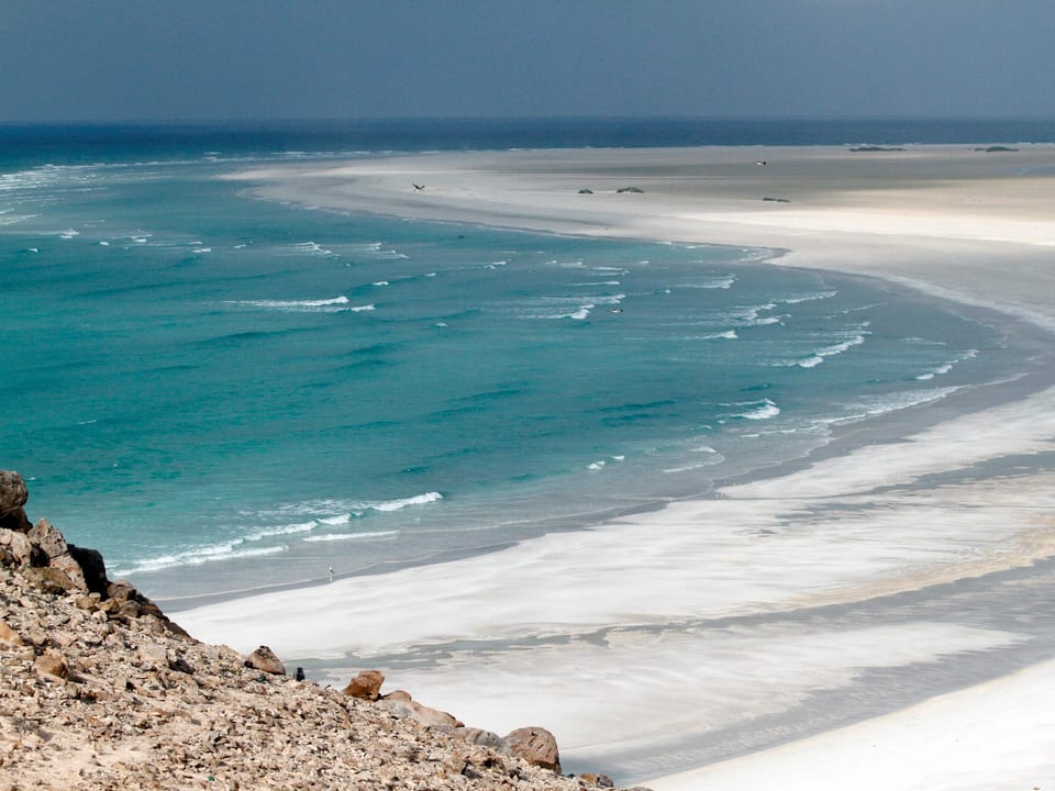 Ein hellgrauer Sandstrand mit leuchtend blauem Meer.