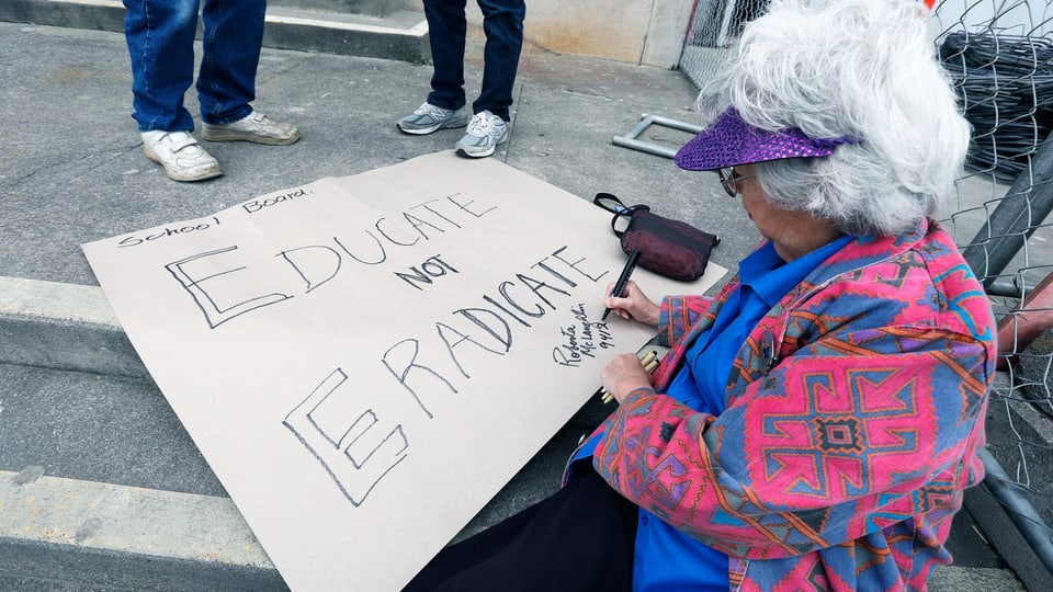 Eine ältere Dame malt ein Transparent. Darauf steht: Educate not eradicate.