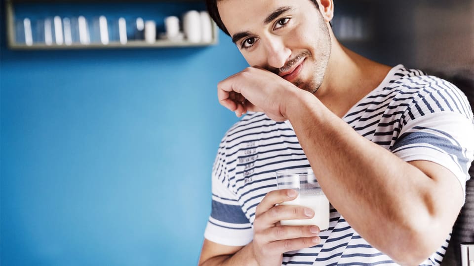 Muskulöser junger Mann hält ein Glas Milch und wischt sich mit der anderen Hand den Milchbart ab.