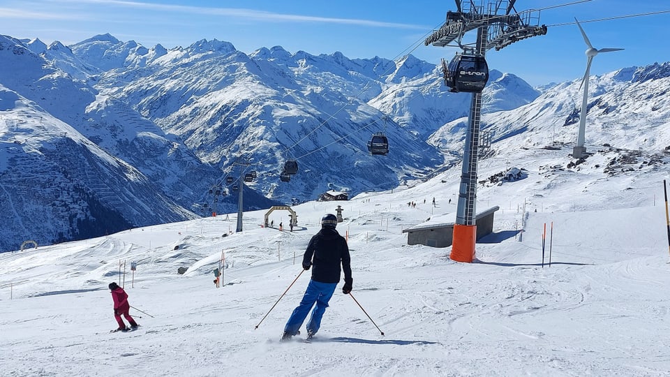 Vorne ein Skifahrer am Fahren auf Piste, hinten Bergpanorama, rechts eine Gondel nahe Mast.