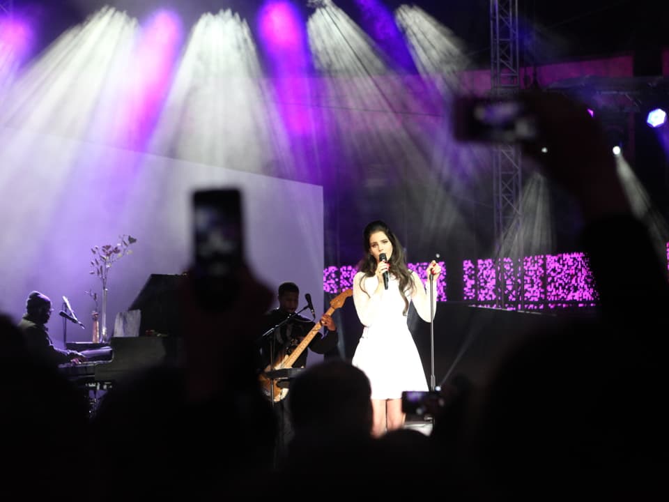 Die Sängerin Lana Del Rey in weissem Kleid und ihre Musiker auf der lila beleuchteten Bühne.