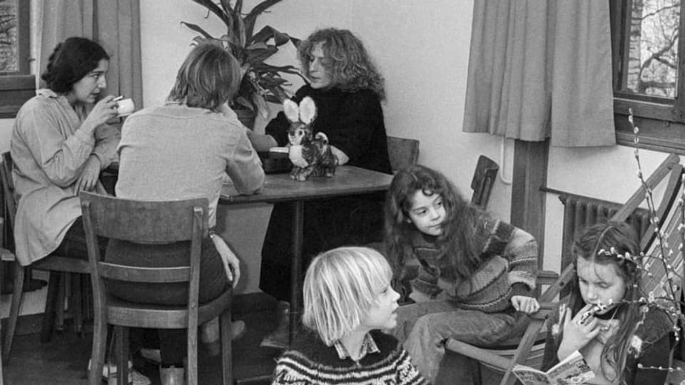 Drei Frauen sitzen an einem runden Tisch. Vor ihnen sind drei Kinder zu sehen.