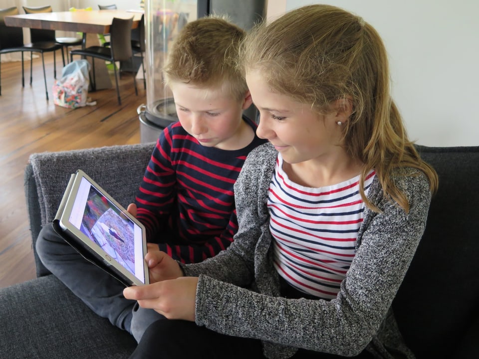 Zwei Kinder sitzen auf einem Sofa und blicken auf ein Tablet.