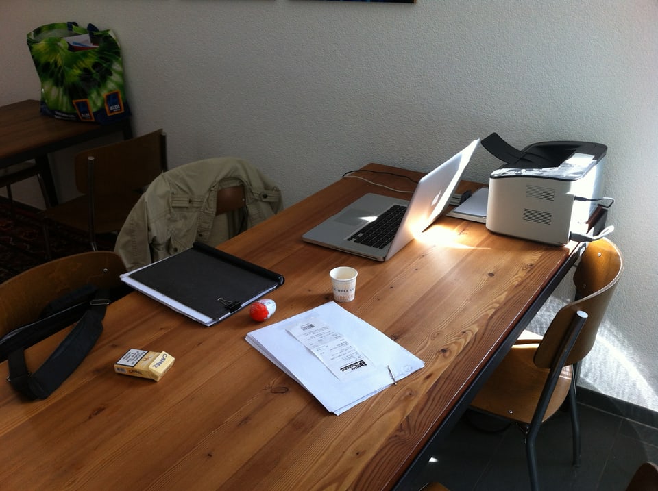 Ein Schreibtisch mit Laptop, Blättern, einem Drucker, einer Packung Zigaretten und einer Mappe.