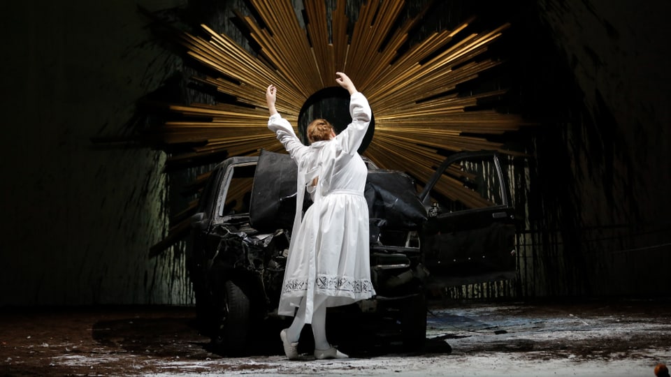 Eine Frau mit weissem Kleid steht auf einer Bühne. Hinter ihr ein Heiligenschein und ein zerstörtes Auto
