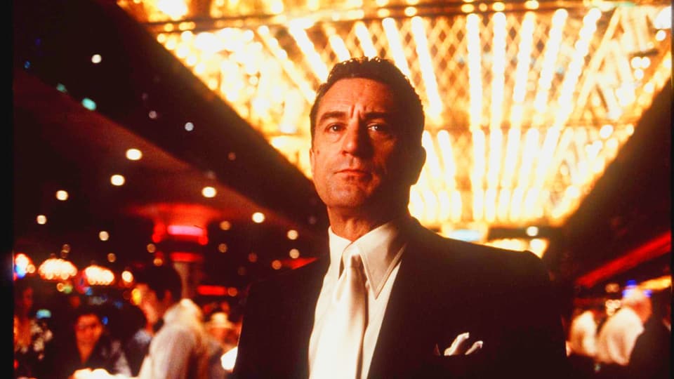 Ein Mann mit gegeeltem Haar in Anzug steht in einem Casino und schaut streng.