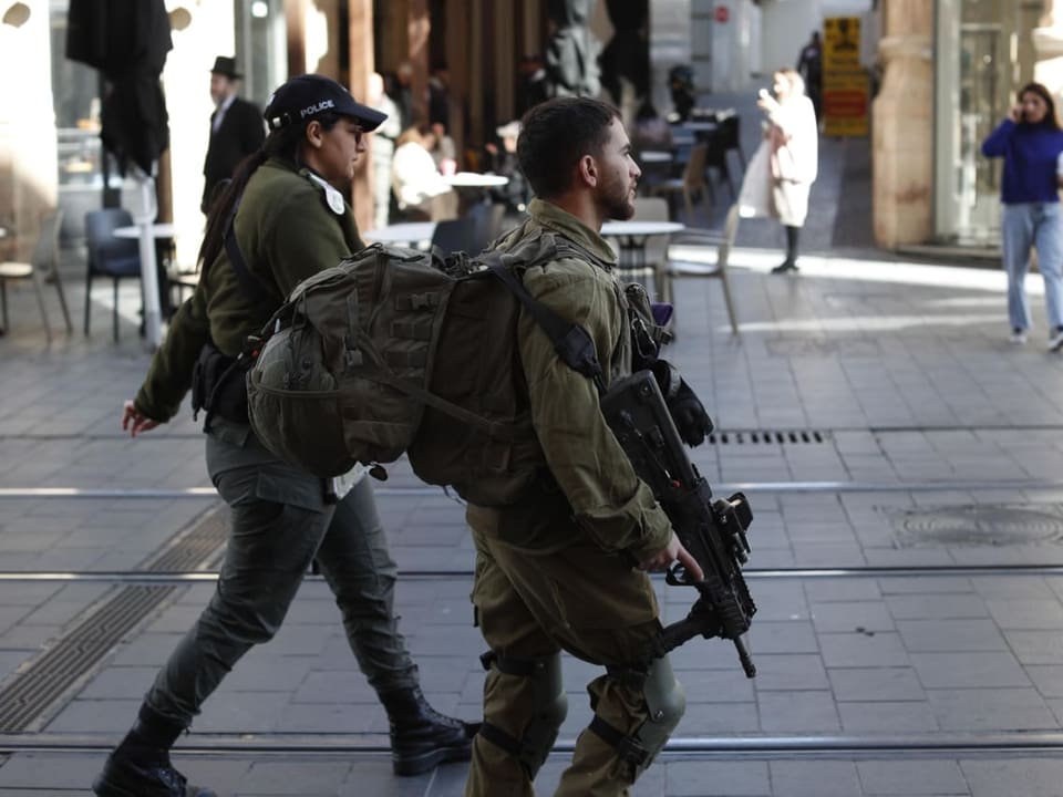 Ein Soldat läuft mit einem Gewehr in einer Fussgängerzone mit Passanten. Neben ihm läuft eine Polizistin.