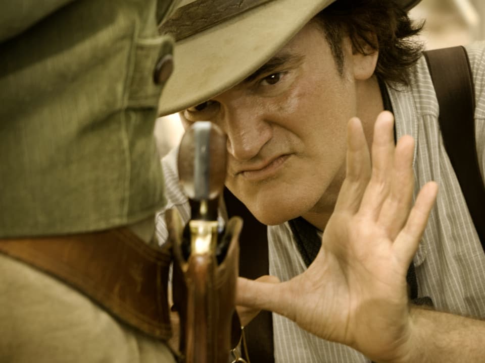 Quentin Tarantino zeigt mit der Hand auf eine Pistole, als solle jemand eine Nahaufnahme davon machen.